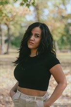 Cheerful hispanic young sensual Woman in a black shirt and beige pants outdoors with hands on hips