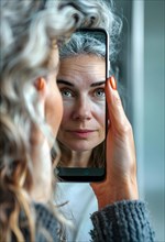 A grey-haired middle-aged woman holds her smartphone in her right hand and looks at her reflection