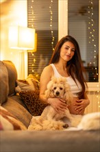 Vertical portrait of a proud pregnant woman and dog sitting together on the sofa at night