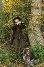 Huntress with hunting dog Griffon, binoculars and rifle at the edge of the forest, Allgaeu,