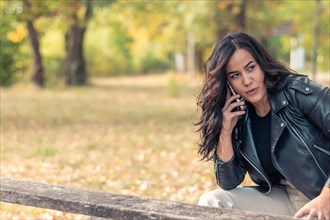 Cheerful hispanic young Woman engaged in a phone call while sitting on a park bench in autumn,