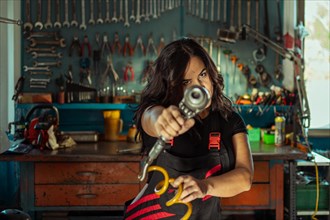 Intense sexy woman mechanic aiming a tool at the camera in a workshop, a complete tool panel in