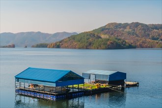 A floating structure with a blue roof on a calm lake with mountains in the distance, in South Korea