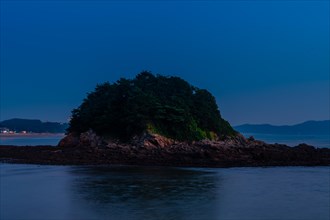 An illuminated island with lush trees stands out against the calm sea during blue hour, in South