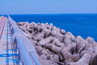 Concrete tetrapods line a coastal walkway under clear skies, with a vivid blue ocean in the