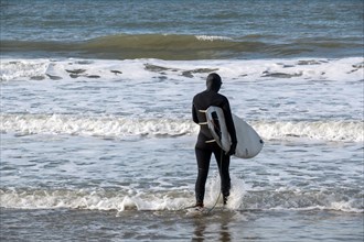 A surfer in a wetsuit goes into the sea with his surfboard
