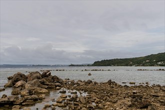 Seascape of rocky shoreline on a cloudy day with buildings on tree lined shore in the distance in