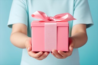 Child's hands holding pink Mother's day gift box with ribbon in front of pastel blue studio