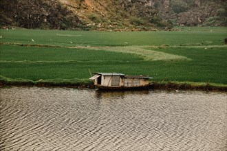 A small boat floats on a river with lush green fields and hills in the background, Ninh Binh,