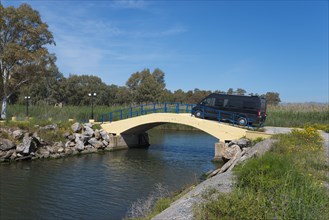 A motorhome drives over a small yellow bridge in a natural landscape on a sunny day, Bouka Beach,