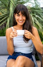 A woman is lookintg at camera while sitting on a couch and holding a white cup. She is smiling and
