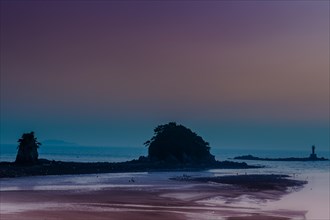 Silhouetted rock formations on a tranquil beach at dawn with a purple sky, in South Korea