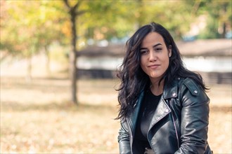 A cheerful hispanic young woman standing in a park with trees wearing a black rider leather jacket,