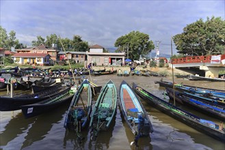 View of calm waterway with anchored boats, a bridge in the background and surrounding houses, Inle