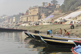 Lively scene by the river where boats lie on the banks and people walk on the ghats by the