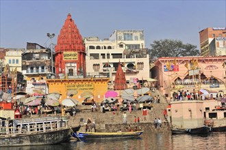 Red temples dominate the ghats by the river, surrounded by people and umbrellas, Varanasi, Uttar