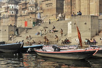 Boats on the bank of a river with people on stairs next to historical buildings, Varanasi, Uttar