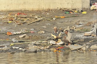 Two people sitting on the polluted bank of a river and seem to be engaged in a conversation,