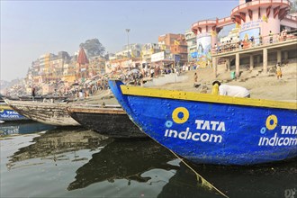Boats on the banks of a busy river with colourful buildings in the background, Varanasi, Uttar