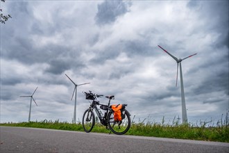 A parked bicycle at the roadside with wind turbines in the background under a cloudy sky, Leer,