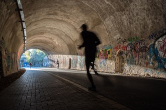 A jogger moves through a tunnel with graffiti and dynamic shadows, Nordbahntrasse, Elberfeld,