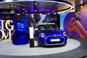 A blue Mini Cooper is presented at a car show next to a large model head, BMW WELT, Munich,
