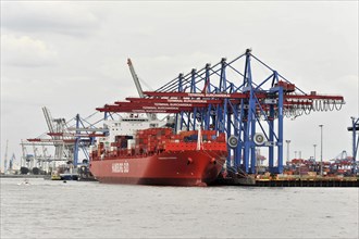 A red container ship in the harbour at a container terminal with numerous cranes, Hamburg,