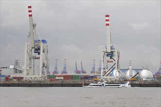 A small boat sails in front of a harbour with large cranes under a cloudy sky, Hamburg, Hanseatic