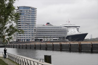 Cruise ship Queen Mary 2, at the dock with modern building in the background under an overcast sky,