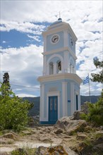 A blue and white clock tower under a sunny sky with the Greek flag and surrounding nature, Poros,