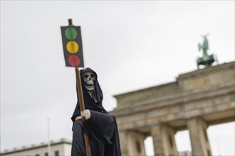 The figure of a grim reaper (skeleton) with a traffic light as a sign, the Brandenburg Gate in the