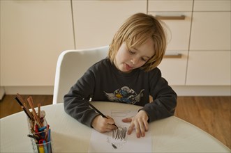Indoor photo, boy, 4-5 years, blond, drawing on sheet of paper, pencils, table, straining, playing