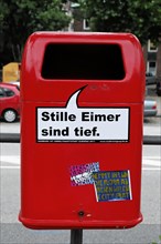 A bright red rubbish bin with stickers in front of a blurred city background, Hamburg, Hanseatic
