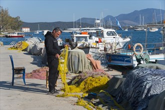 A man repairs traditional fishing nets at the sunny marina with boats and mountains in the
