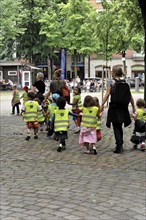 Group of children with high-visibility waistcoats on the hand of an adult in an urban environment,