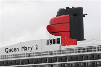The picture shows the smokestack of the Queen Mary 2, white and red, in front of a cloudy sky,