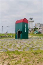 Green post box in a field with a lighthouse in the background under a cloudy sky, in Ulsan, South