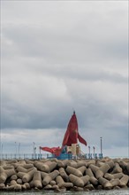 A red cloth flutters in the wind on a breaker with concrete blocks against a cloudy ocean backdrop,