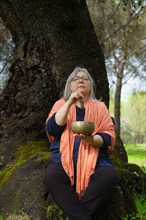 Mature woman with white hair in her sixties meditating in the forest with her eyes closed and a