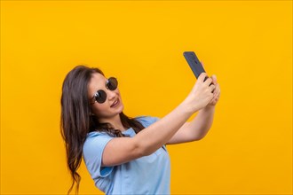 Studio portrait with yellow background of a casual woman with sunglasses taking a selfie with