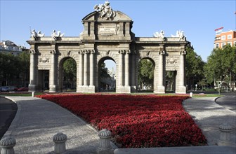 Historical monument with arches and sculptures, surrounded by a red flowerbed under a clear sky
