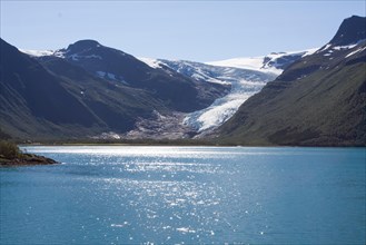 A glacier flows between snow-capped mountains into a clear blue lake under bright sunshine Arctic