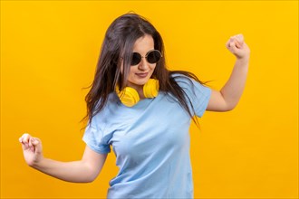 Studio portrait with yellow background of happy woman dancing wearing sunglasses and headphones on
