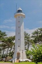 A tall white lighthouse tower against a clear blue sky surrounded by trees, in Ulsan, South Korea,
