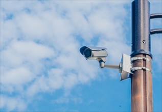 Surveillance camera mounted on metal light pole with blue sky and puffy white clouds in background