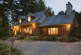 Illuminated old 1800s reconstructed log cabin home facade and paving stone driveway at dusk,
