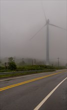Large electric wind turbine hidden by heavy morning fog next to a two lane highway in countryside