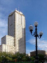A skyscraper rises into the blue sky, next to a classic street lamp and surrounded by greenery