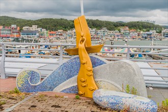 A colorful mosaic fish sculpture at a harbor with boats and a cloudy sky in the background, in