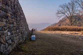 Park bench beside mountain fortress wall made of flat stones with hazy blue sky in background in
